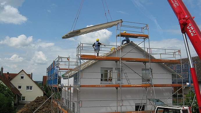 Symbolfoto Kran Bauelement schwebt in der Luft, zwei Bauarbeiter auf Dach